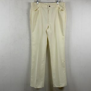 May include: Cream-colored, straight-leg pants hanging on a metal hanger. The pants have a button closure and two front pockets. The fabric appears to be a lightweight material. The pants are hanging against a white paneled wall.