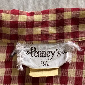 May include: A white tag with the text "Penney's" and the size "13/14" on a red and yellow gingham fabric.