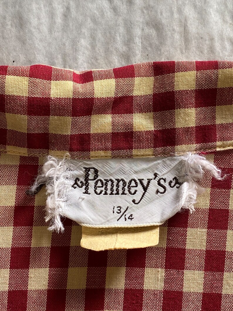 May include: A white tag with the text "Penney's" and the size "13/14" on a red and yellow gingham fabric.