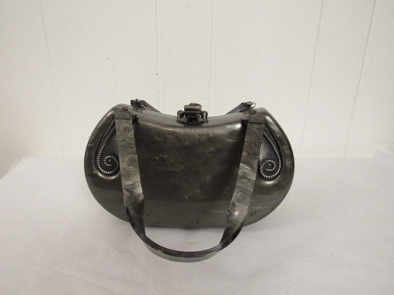 May include: A silver-colored, oval-shaped clutch purse with a decorative clasp and a black strap.