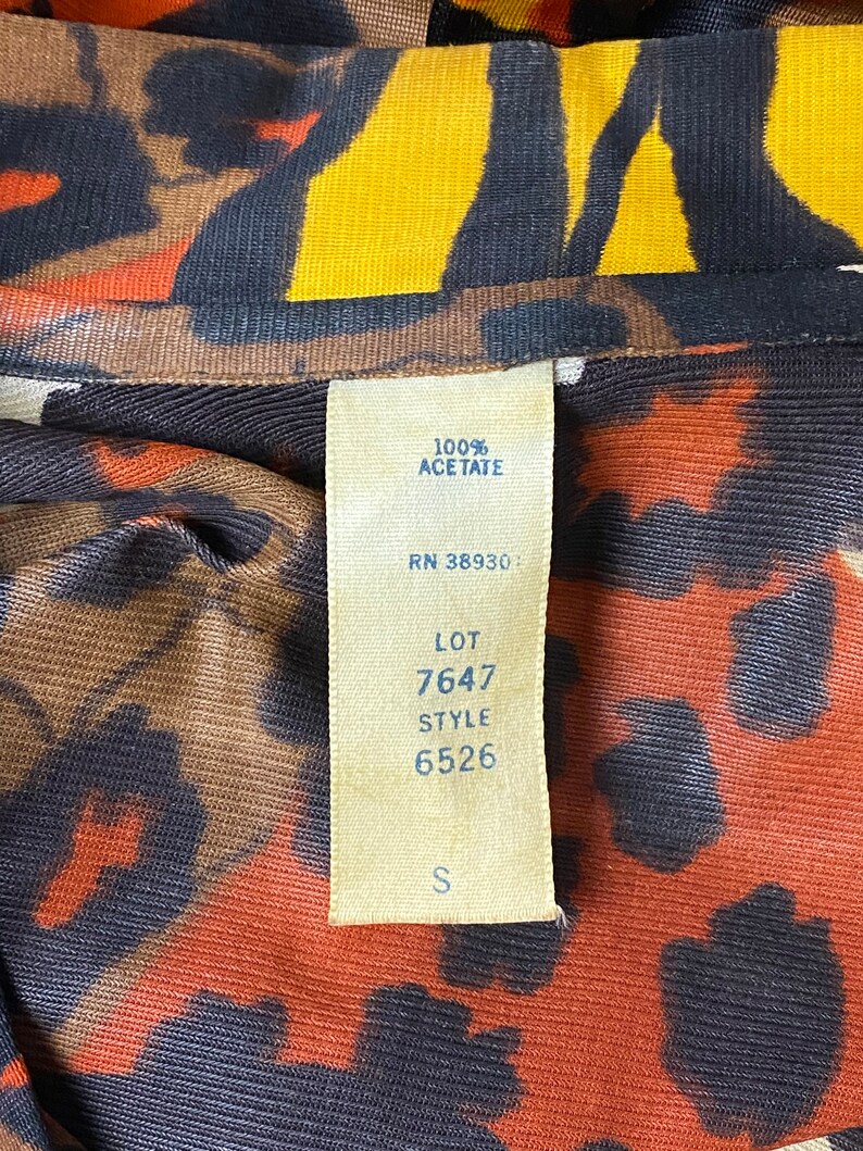 Puede incluir: Una tela de estampado de leopardo marr&oacute;n, naranja y amarillo con una etiqueta blanca adjunta. La etiqueta tiene el texto "100% Acetato", "RN 38930", "LOT 7647", "STYLE 6526" y "S".