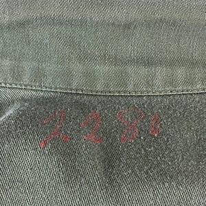 May include: Close-up of a section of olive green fabric, possibly denim or canvas. The number "2281" is hand-stitched in red thread near the collar. The fabric has a textured appearance.