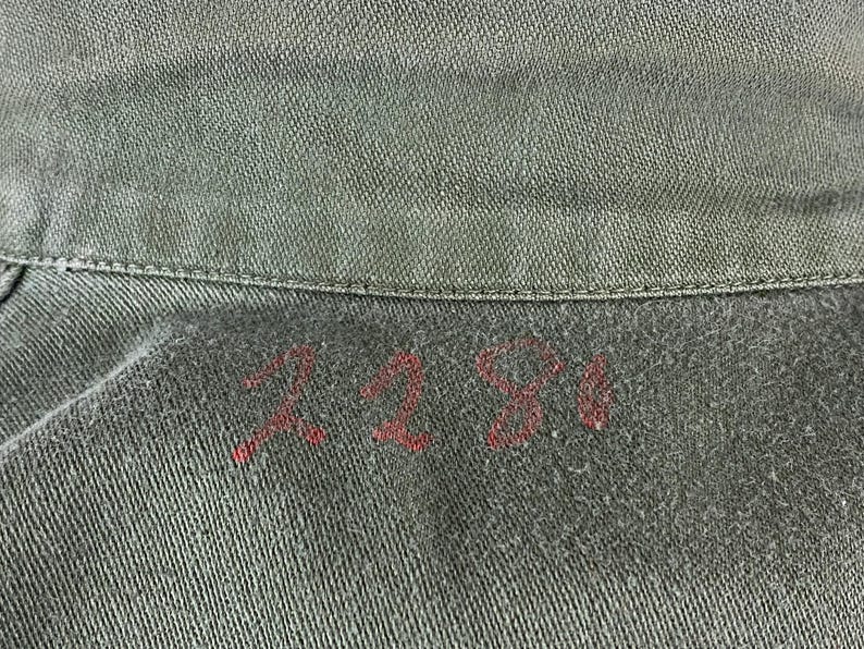 May include: Close-up of a section of olive green fabric, possibly denim or canvas. The number "2281" is hand-stitched in red thread near the collar. The fabric has a textured appearance.