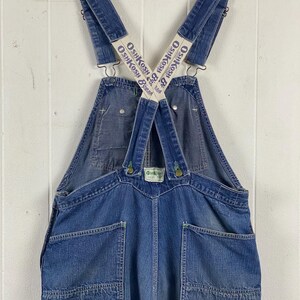 K&ouml;nnte beinhalten: Vintage blaue Jeans-Latzhose mit dem OshKosh B'gosh-Logo auf den Tr&auml;gern. Die Latzhose hat mehrere Taschen, Metallbeschl&auml;ge und ein klassisches Latzdesign. Das Kleidungsst&uuml;ck h&auml;ngt an einem Metallb&uuml;gel vor einem wei&szlig;en Hintergrund.