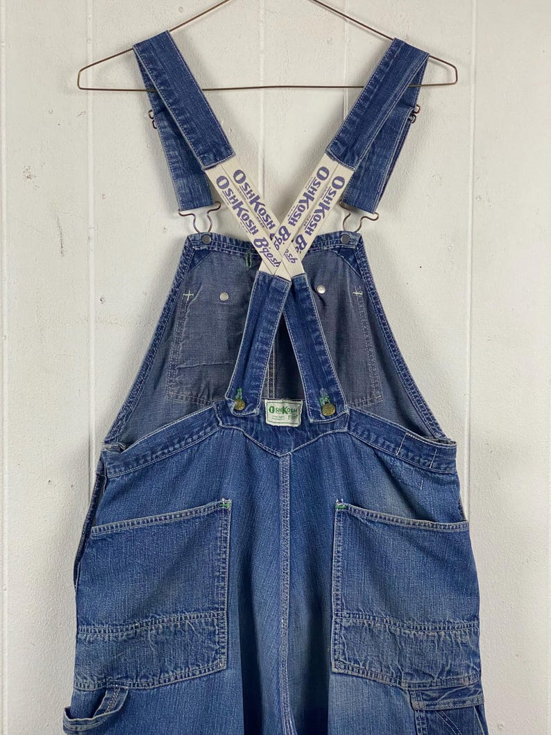K&ouml;nnte beinhalten: Vintage blaue Jeans-Latzhose mit dem OshKosh B'gosh-Logo auf den Tr&auml;gern. Die Latzhose hat mehrere Taschen, Metallbeschl&auml;ge und ein klassisches Latzdesign. Das Kleidungsst&uuml;ck h&auml;ngt an einem Metallb&uuml;gel vor einem wei&szlig;en Hintergrund.
