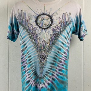 Puede incluir: Una camiseta tie-dye con un dise&ntilde;o de cristal colorido y un planeta en el centro. La camiseta es de color azul claro, blanco y morado.