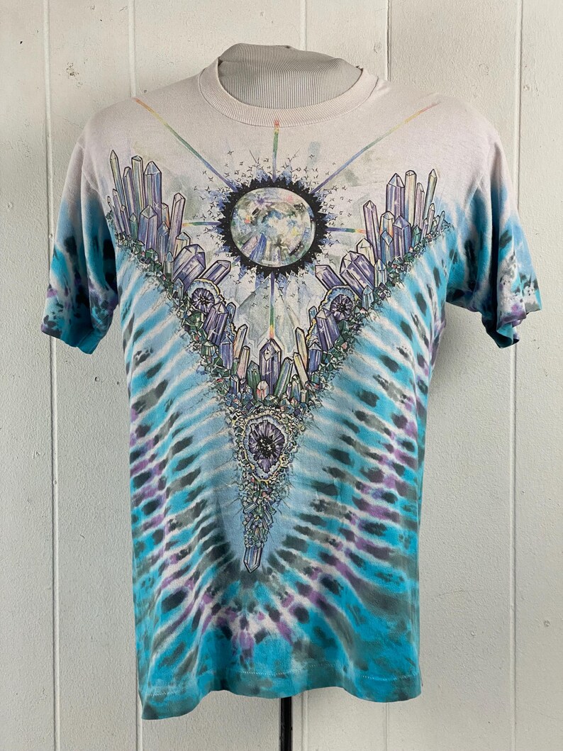 Puede incluir: Una camiseta tie-dye con un dise&ntilde;o de cristal colorido y un planeta en el centro. La camiseta es de color azul claro, blanco y morado.