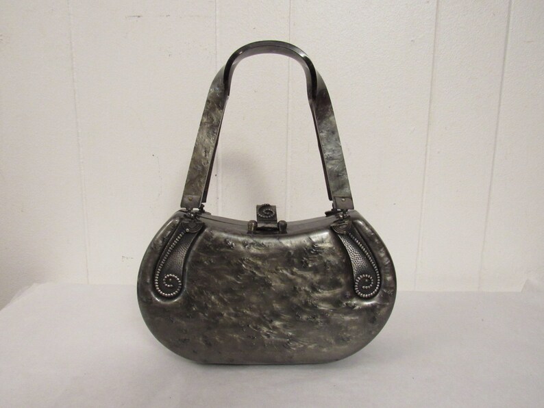 May include: A silver-colored, oval-shaped handbag with a black plastic handle. The bag has a textured surface and decorative details on the sides.