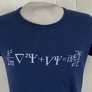 Könnte beinhalten: Marineblaues T-Shirt mit einer weißen mathematischen Gleichung, wahrscheinlich aus der Quantenmechanik. Die Gleichung ist in einer serifenlosen Schrift gedruckt. Das T-Shirt hat einen Rundhalsausschnitt und ist aus weichem Material.