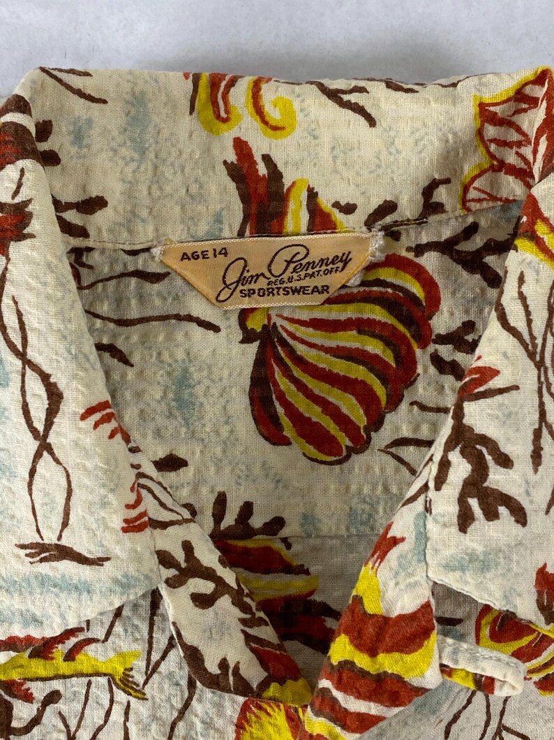 May include: A vintage beige button-up shirt with a colorful floral print. The shirt has a label that reads "Jim Penny Sportswear" and "Age 14".