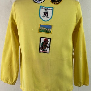 May include: A yellow long-sleeved shirt with various embroidered patches. The patches include "Amana Colonies in Iowa", "Massachusetts", "Hollywood", "Gemini", and "I'm a Rockhound".