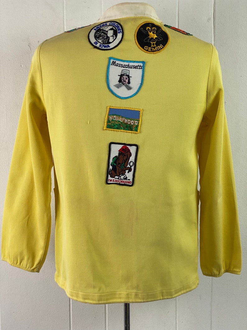 May include: A yellow long-sleeved shirt with various embroidered patches. The patches include "Amana Colonies in Iowa", "Massachusetts", "Hollywood", "Gemini", and "I'm a Rockhound".
