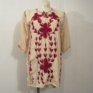 May include: A cream-colored tunic with red embroidered floral designs. The tunic has a round neckline and three-quarter sleeves.