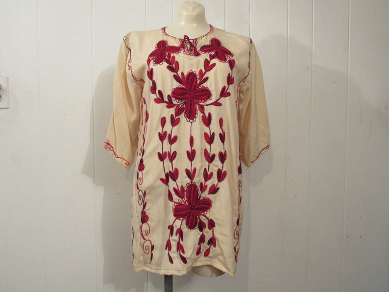 May include: A cream-colored tunic with red embroidered floral designs. The tunic has a round neckline and three-quarter sleeves.
