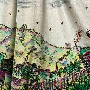 May include: A white skirt with a colorful printed design of a city skyline with mountains in the background. The skirt is decorated with small, colorful beads.