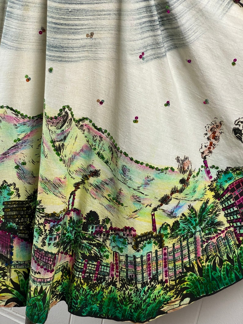 May include: A white skirt with a colorful printed design of a city skyline with mountains in the background. The skirt is decorated with small, colorful beads.