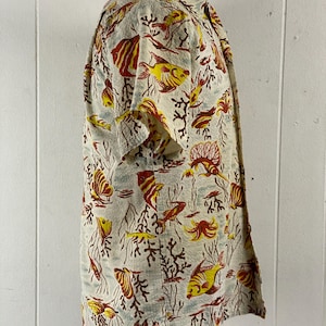 May include: A short-sleeved button-up shirt with a white background and a colorful print of fish and coral. The shirt has a relaxed fit and a classic collar.