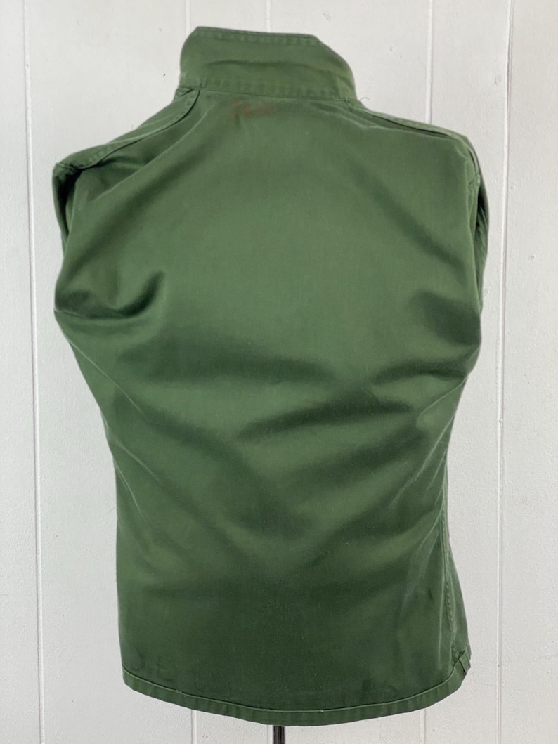 May include: A solid olive green jacket with a high collar. The back of the jacket is visible, showing the tailored fit and seams. The fabric appears to be a durable, possibly military-style material.