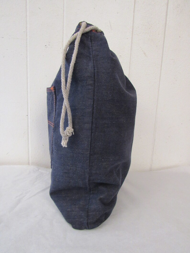 K&ouml;nnte beinhalten: Eine dunkelblaue Kordeltasche aus Denim mit einer kleinen Tasche an der Seite. Die Tasche ist aus einem dicken, strapazierf&auml;higen Stoff gefertigt und hat einen wei&szlig;en Kordelzug.