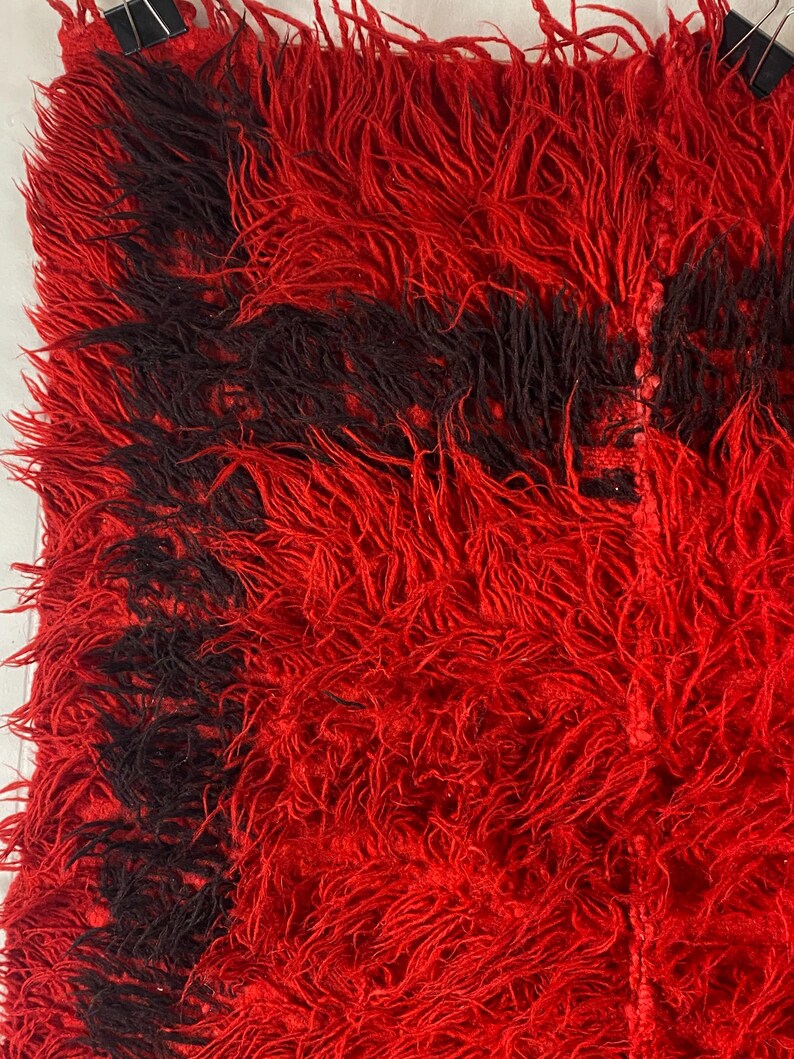 May include: Red and black shaggy rug with a fringe border. The rug has a unique texture and is made of soft, fluffy fibers.