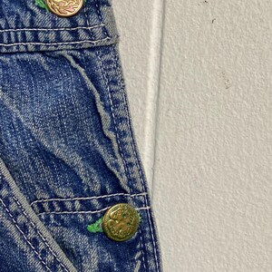 K&ouml;nnte beinhalten: Nahaufnahme einer blauen Jeans-Latzhose mit goldfarbenen Kn&ouml;pfen. Die Kn&ouml;pfe haben ein dekoratives Design und sind mit gr&uuml;nem Faden befestigt. Wei&szlig;e N&auml;hte akzentuieren die N&auml;hte des Jeansstoffs.