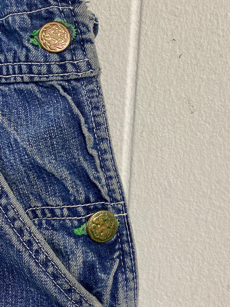 K&ouml;nnte beinhalten: Nahaufnahme einer blauen Jeans-Latzhose mit goldfarbenen Kn&ouml;pfen. Die Kn&ouml;pfe haben ein dekoratives Design und sind mit gr&uuml;nem Faden befestigt. Wei&szlig;e N&auml;hte akzentuieren die N&auml;hte des Jeansstoffs.