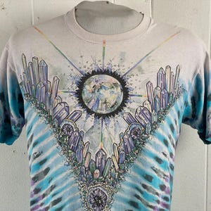 Puede incluir: Una camiseta blanca tie-dye con un dise&ntilde;o de cristal y tierra colorido. La camiseta presenta un gran dise&ntilde;o de cristal colorido sobre un fondo tie-dye azul y blanco.