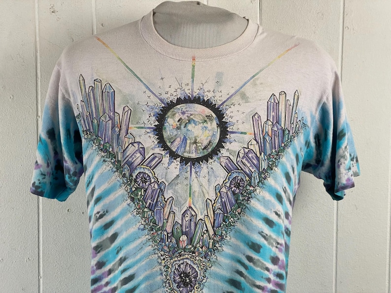 Puede incluir: Una camiseta blanca tie-dye con un dise&ntilde;o de cristal y tierra colorido. La camiseta presenta un gran dise&ntilde;o de cristal colorido sobre un fondo tie-dye azul y blanco.