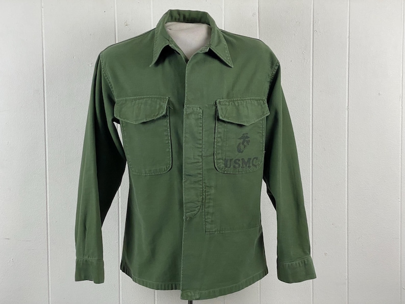 May include: Olive green military-style shirt with two chest pockets and long sleeves. The shirt features a collar and a button-up front. The USMC emblem and text are visible on the right chest. The shirt is displayed on a mannequin.