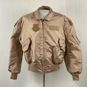 May include: Tan bomber jacket with multiple pockets, a ribbed collar, and cuffs. The jacket has patches on the chest and sleeves. The jacket is made of a shiny, nylon-like material.