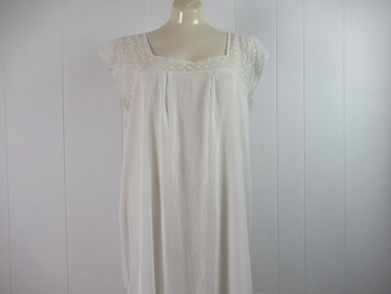 Puede incluir: Un camis&oacute;n blanco de algod&oacute;n con ribete de encaje alrededor del escote y las mangas. El camis&oacute;n tiene un escote cuadrado y un corte holgado.