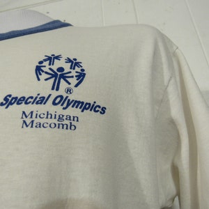 May include: White long-sleeve t-shirt with a blue Special Olympics logo featuring a circle of stick figures holding hands. The text "Special Olympics" is written in blue below the logo, followed by "Michigan Macomb" in smaller blue text.