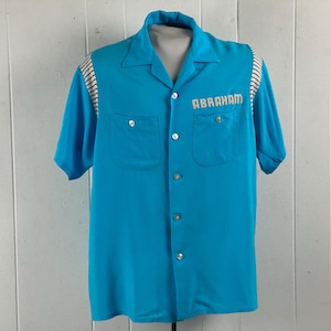 Puede incluir: Una camisa de botones de manga corta de color azul turquesa con rayas blancas en los hombros y la palabra "ABRAHAM" bordada en la parte delantera.