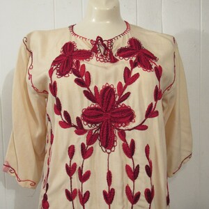May include: A cream-colored tunic with red embroidered floral designs. The embroidery features a large flower in the center and smaller flowers and leaves around it.