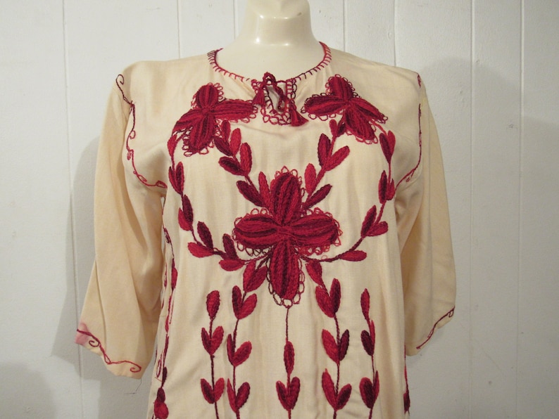 May include: A cream-colored tunic with red embroidered floral designs. The embroidery features a large flower in the center and smaller flowers and leaves around it.