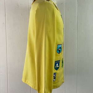 May include: A yellow jacket with a variety of colorful patches sewn on the front. The patches feature images of birds, flowers, and other nature-themed designs.