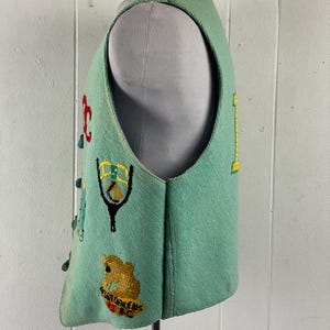 May include: A teal blue vest with colorful embroidered patches. The patches include a slingshot, a bear, and the text "SUGAR CREEK CAPS FC".