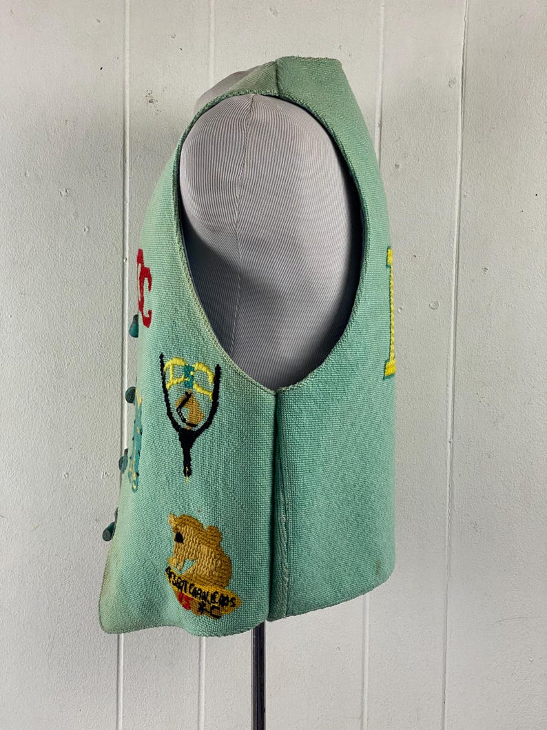 May include: A teal blue vest with colorful embroidered patches. The patches include a slingshot, a bear, and the text "SUGAR CREEK CAPS FC".