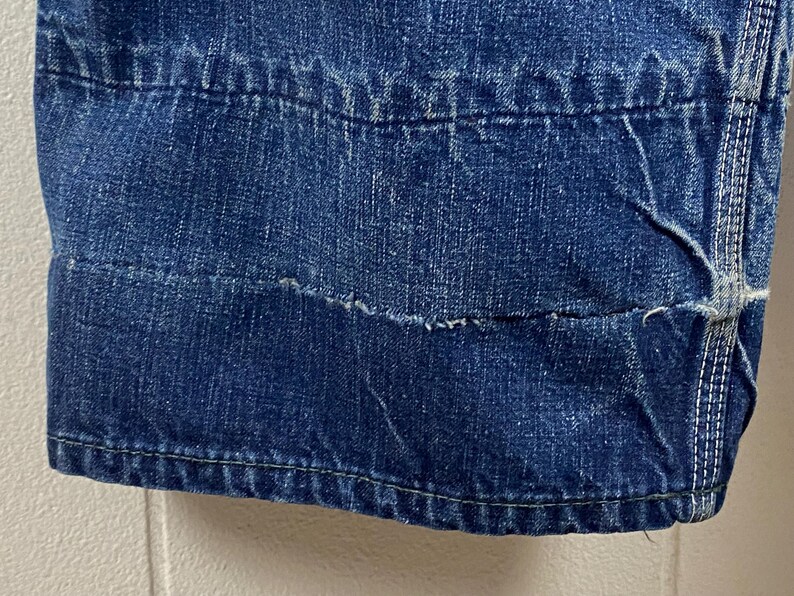 K&ouml;nnte beinhalten: Nahaufnahme eines Abschnitts einer blauen Jeans. Das Bild zeigt die Nahtdetails, einschlie&szlig;lich der N&auml;hte und der verst&auml;rkten Bereiche. Der Denim hat ein abgenutztes Aussehen mit sichtbaren Gebrauchsspuren und einem ausgefransten Rand.