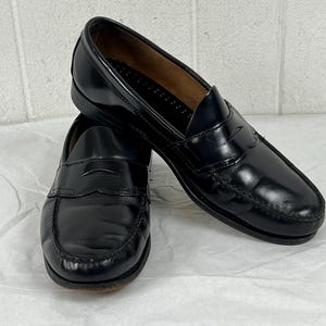 May include: A pair of black leather penny loafers featuring a classic moc-toe construction, a horizontal strap across the vamp with a signature cutout, a low stacked heel, and a smooth polished finish with visible stitching along the rounded toe.