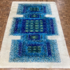 May include: A rectangular shag rug with a geometric design. The rug features a central pattern of blue and green rectangles on a white background. The rug's texture appears soft and plush, suitable for a living room or bedroom.