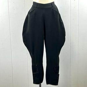Puede incluir: Pantalones de equitación negros lisos con diseño de cintura alta, paneles laterales ensanchados en las caderas y secciones inferiores reforzadas en las piernas con cierres de botones verticales en los tobillos, sobre un maniquí.