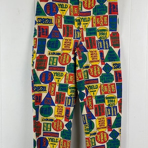 Könnte beinhalten: Eine cremefarbene Hose mit einem bunten Muster aus Schildern und Sätzen. Die Schilder sind in verschiedenen Formen und Farben, darunter Rot, Gelb, Blau und Grün. Einige der Sätze auf den Schildern lauten "Vorfahrt gewähren", "Power", "Angelurlaub", "Vogelbäder sind unanständig" und "Liebe verbreitet Keime".