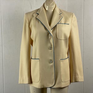 May include: A cream-colored blazer with blue trim around the edges of the lapels, pockets, and button placket. The blazer has three buttons and two pockets.
