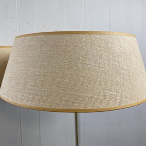 May include: Two beige lampshades with a textured fabric and a light brown trim. The lampshades have a classic, conical shape and are set against a light grey background.
