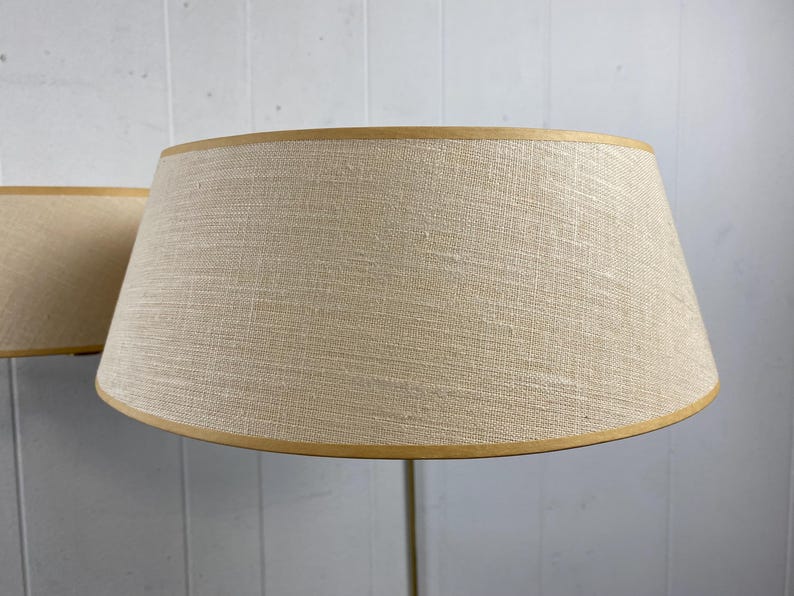 May include: Two beige lampshades with a textured fabric and a light brown trim. The lampshades have a classic, conical shape and are set against a light grey background.