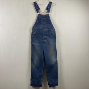 K&ouml;nnte beinhalten: Blaue Jeans-Latzhose an einem Metallb&uuml;gel. Die Latzhose hat verstellbare Tr&auml;ger, Knopfverschl&uuml;sse und Taschen. Der Denim weist Gebrauchsspuren auf, mit Verf&auml;rbungen und Ausbleichen. Das Kleidungsst&uuml;ck wird vor einer wei&szlig;en Wand mit Paneelen pr&auml;sentiert.