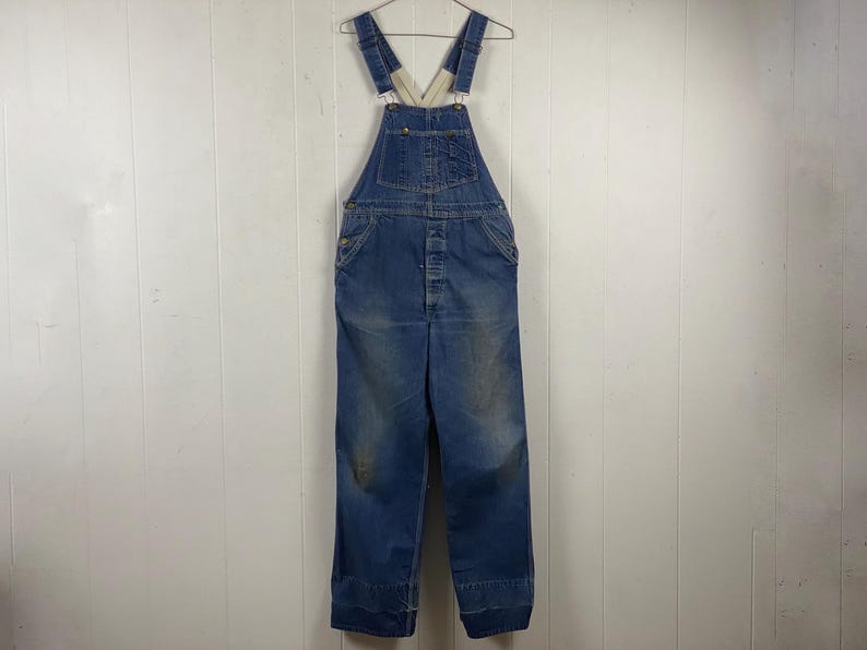 K&ouml;nnte beinhalten: Blaue Jeans-Latzhose an einem Metallb&uuml;gel. Die Latzhose hat verstellbare Tr&auml;ger, Knopfverschl&uuml;sse und Taschen. Der Denim weist Gebrauchsspuren auf, mit Verf&auml;rbungen und Ausbleichen. Das Kleidungsst&uuml;ck wird vor einer wei&szlig;en Wand mit Paneelen pr&auml;sentiert.