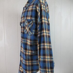 以下が含まれることがあります： 青、黄、茶色の格子柄のフランネルシャツ。長袖でボタンダウンフロントです。