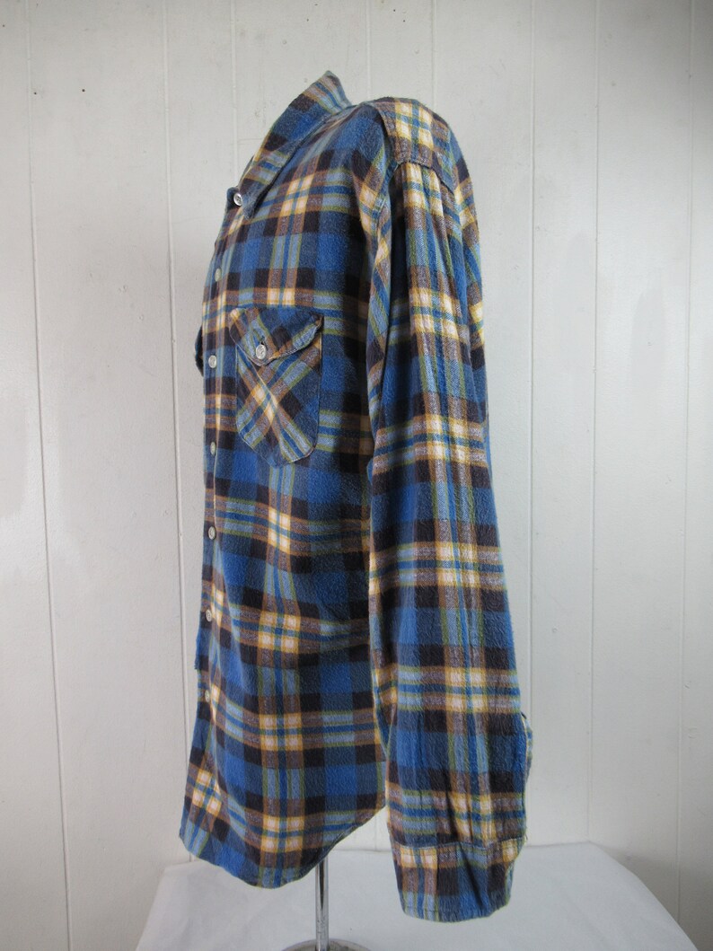 以下が含まれることがあります： 青、黄、茶色の格子柄のフランネルシャツ。長袖でボタンダウンフロントです。
